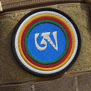 Puede incluir: Un parche bordado circular con un centro azul y un carácter de escritura tibetana en blanco, rodeado por anillos concéntricos de color verde, rojo, amarillo, blanco y negro, sujeto a una mochila táctica de color tostado con cremallera.