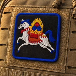Può includere: Una toppa ricamata quadrata raffigurante un cavallo bianco con criniera e coda blu, che trasporta una sella gialla e rossa fiammeggiante, su sfondo nero con bordo blu, fissata a uno zaino tattico color marrone chiaro con fettuccia MOLLE.