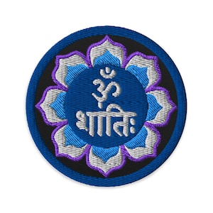 Peut inclure: Écusson brodé rond avec un cercle bleu central orné du symbole Om et du mot "Shanti" en blanc. Le motif comprend une fleur de lotus avec des pétales bleus, blancs et violets, sur fond noir.