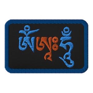 Può includere: Una toppa ricamata blu e arancione con la frase tibetana "Om Mani Padme Hum" su uno sfondo nero.