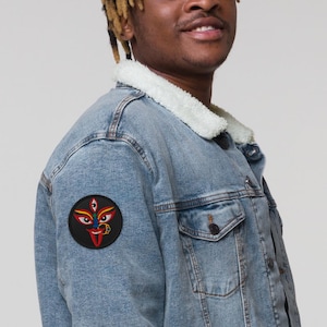 May include: Light blue denim jacket with a cream-colored sherpa collar. A round patch featuring a colorful face design with three eyes, a red tongue, and gold jewelry is sewn onto the jacket's sleeve. The jacket has a button-down closure and two chest pockets.