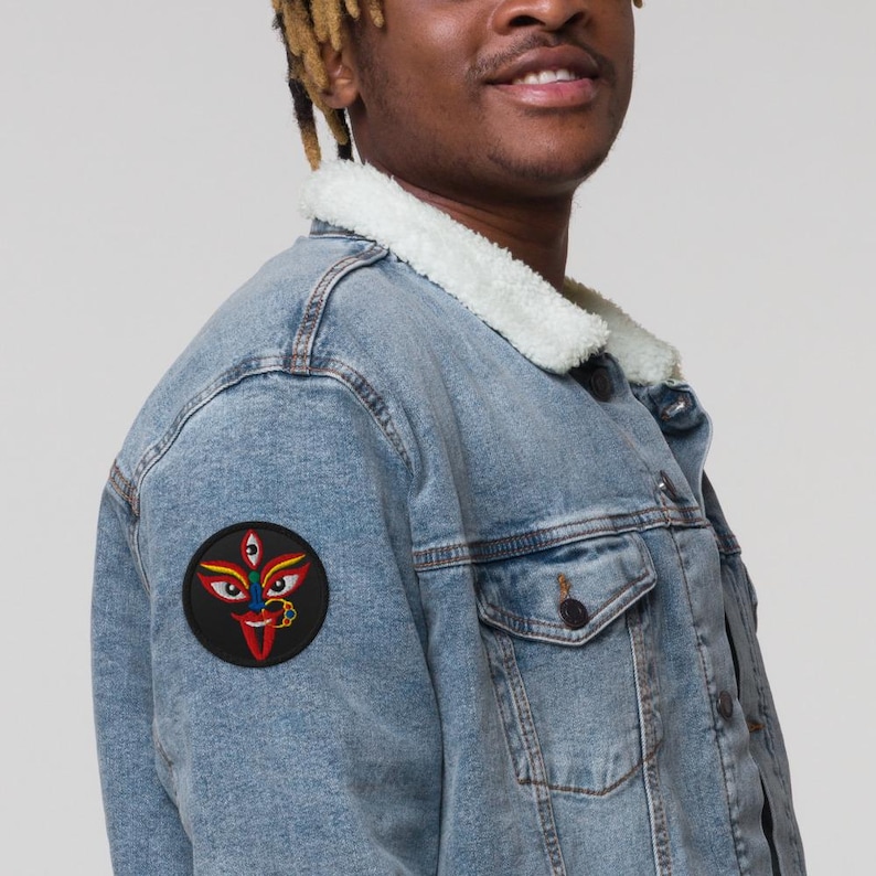 May include: Light blue denim jacket with a cream-colored sherpa collar. A round patch featuring a colorful face design with three eyes, a red tongue, and gold jewelry is sewn onto the jacket's sleeve. The jacket has a button-down closure and two chest pockets.