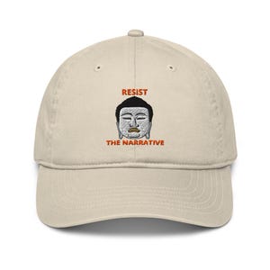 Op de afbeelding: Een beige baseballpet met een geborduurd beeld van een Boeddha met de tekst "Resist the Narrative" in oranje.