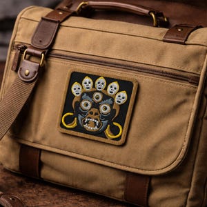 May include: A tan canvas messenger bag with brown leather straps and a rectangular embroidered patch featuring a black, white, and yellow stylized face with three eyes, bared teeth, and a crown of five small skulls on a black background.
