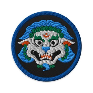 Puede incluir: Parche bordado con la cabeza estilizada de un león con melena y detalles azules. El león tiene la cara blanca, orejas naranjas y nariz azul. El parche es redondo con fondo negro y borde azul.