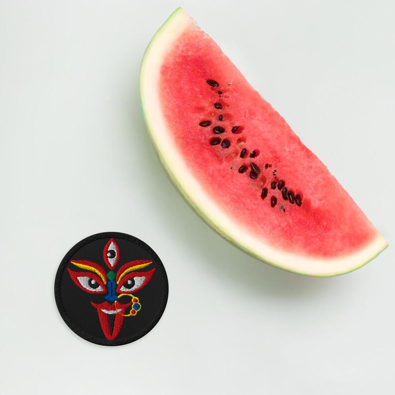 May include: A black embroidered patch featuring a colorful depiction of a deity's face with three eyes, red lips, and a nose ring. A slice of watermelon with black seeds is also in the image.