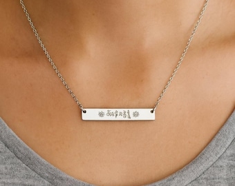 May include: A silver-toned horizontal bar necklace featuring a delicate chain and a rectangular pendant. The pendant is engraved with a series of intricate, small symbols and characters centered across its polished, flat metallic surface.