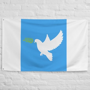 May include: A white flag with a central blue stripe featuring a white dove carrying an olive branch. The flag has white borders on the left and right sides, and is hanging against a white brick wall.