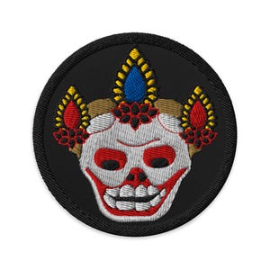 Puede incluir: Parche bordado con una calavera estilizada con una amplia sonrisa y ojos rojos. La calavera es blanca con detalles rojos y negros, rodeada por un borde circular negro. El diseño incluye elementos decorativos amarillos, azules y marrones.