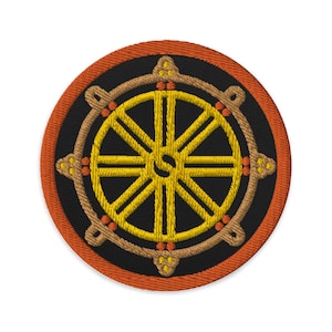 May include: A round embroidered patch with a Dharmachakra, or Wheel of Law, in yellow and brown on a black background. The patch has an orange border.