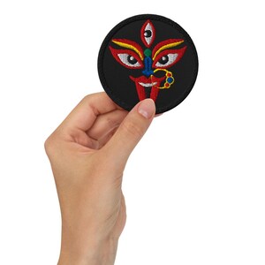 May include: A round, black embroidered patch featuring a stylized face with red, yellow, blue, and green details. The patch is held in a hand, showcasing the intricate design and vibrant colors. The design includes a third eye and a nose ring.