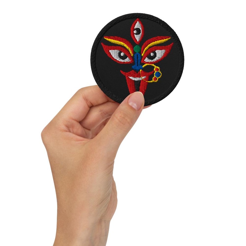 May include: A round, black embroidered patch featuring a stylized face with red, yellow, blue, and green details. The patch is held in a hand, showcasing the intricate design and vibrant colors. The design includes a third eye and a nose ring.