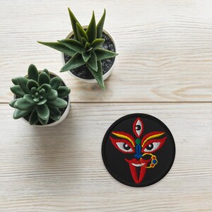 May include: A round, black embroidered patch featuring a stylized face with three eyes, red lips, and yellow and red accents. The patch is on a light wood-grain surface, with two small potted succulents in the background.
