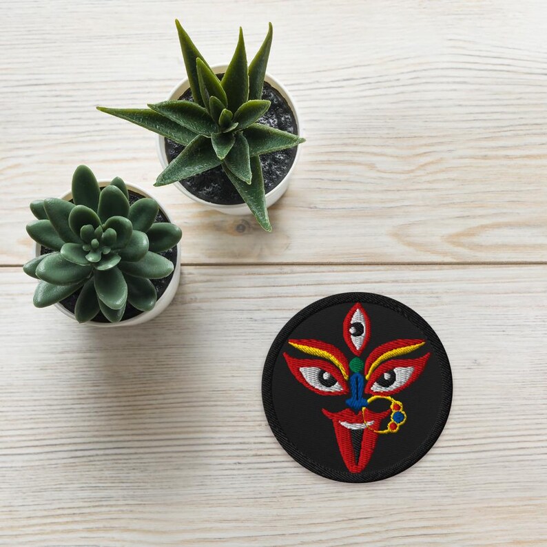 May include: A round, black embroidered patch featuring a stylized face with three eyes, red lips, and yellow and red accents. The patch is on a light wood-grain surface, with two small potted succulents in the background.