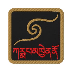 Puede incluir: Un parche bordado en dorado con fondo negro. El parche presenta un diseño de espiral estilizado y el texto "གསང་སྔགས་གླིང་" en rojo.