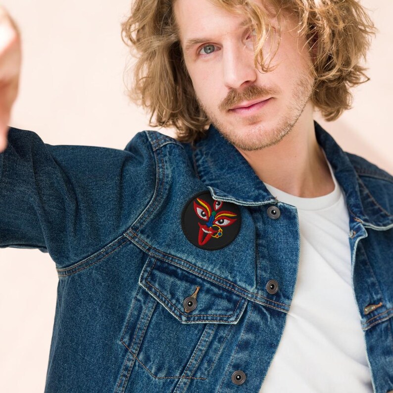 May include: A denim jacket with a round patch featuring a colorful, stylized face with three eyes, red lips, and gold accents. The patch is affixed to the jacket's chest. The jacket is a medium wash blue.