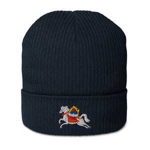 Pode incluir: Gorro azul marinho com textura canelada. O gorro apresenta um desenho bordado de um cavalo branco com uma sela vermelha e um escudo decorativo. O cavalo está em posição de corrida. Um acessório de inverno.