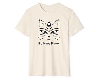 May include: A cream-coloured t-shirt with a black cat face design and the text "Be Here Meow". The cat face has stylized eyes, whiskers, and a third eye. The shirt is a short-sleeved crew neck.