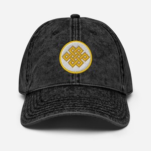 May include: Black baseball cap with a worn look. It has a gold and white embroidered knot symbol on the front. The cap has a curved peak and a button on top. The design is circular.