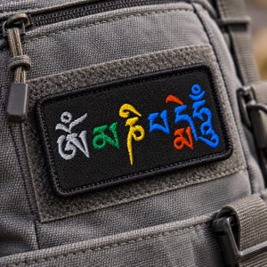 Puede incluir: Un parche rectangular negro con escritura tibetana bordada en hilos de color blanco, verde, amarillo, azul y rojo. El parche está sujeto a una bolsa de lona gris con una cremallera y una lengüeta de tela visibles en la parte superior izquierda.