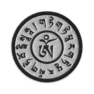 Op de afbeelding: Een geborduurde patch in zwart en wit met een cirkelvormig ontwerp. Het midden heeft een gestileerde figuur met een symbool. De buitenste cirkel heeft een herhalend patroon van symbolen.
