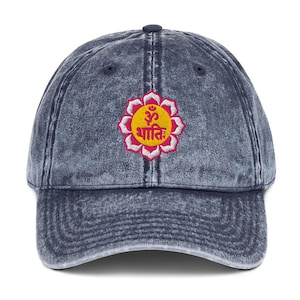 Puede incluir: Una gorra de béisbol de mezclilla azul con un diseño de flor bordado en amarillo y rosa. La flor tiene un símbolo Om en el centro y la palabra "Shanti" escrita en sánscrito.