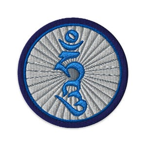 Pode incluir: Um emblema bordado circular com um símbolo tibetano Hum azul brilhante centrado num fundo cinzento prateado com um padrão de raios de sol. O emblema tem um rebordo cosido em azul-marinho escuro em toda a sua circunferência.