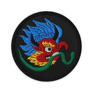 Op de afbeelding: Geborduurde patch met een gestileerde drakenkop. De draak heeft een rood gezicht, gele manen, blauw haar en een groene, slangachtige tong. De patch is cirkelvormig met een zwarte achtergrond en rand.