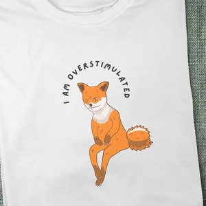 White t-shirt with me fox sitting looking troubled with black, curved text above image that says "I am overstimulated" in capital letters.