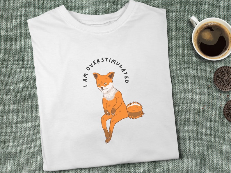 White t-shirt with me fox sitting looking troubled with black, curved text above image that says "I am overstimulated" in capital letters.
