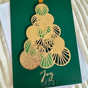May include: A Christmas card with a gold-colored tree design on a dark green background. The tree is made of overlapping circular shapes and topped with a gold star. The word "Joy" is written in gold script.