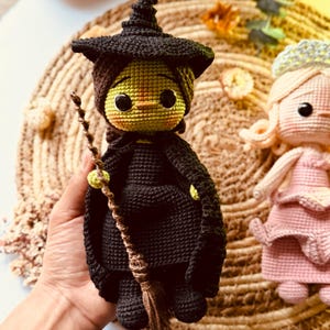 Puede incluir: Muñeca de bruja de ganchillo con sombrero negro, capa y escoba. La muñeca tiene una cara verde, ojos grandes y una cofia marrón. También se ve una segunda muñeca de ganchillo con un vestido rosa.