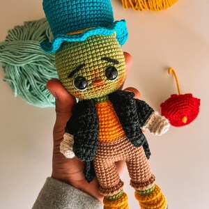 May include: Crocheted doll with a green head, wearing a blue top hat, black jacket, orange shirt, and brown trousers. The doll has large black eyes and is holding a small red umbrella. The doll is approximately 15 cm tall.
