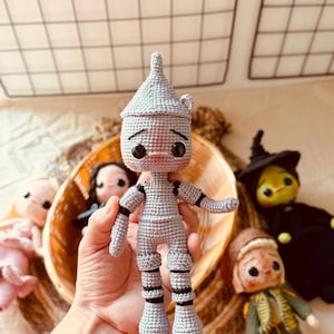 Puede incluir: Muñeco de ganchillo del Hombre de Hojalata en gris con detalles negros, parte de una colección del Mago de Oz. El muñeco tiene un sombrero cónico, ojos negros grandes y mide aproximadamente 15 cm de alto. Otros muñecos de ganchillo son visibles en el fondo.