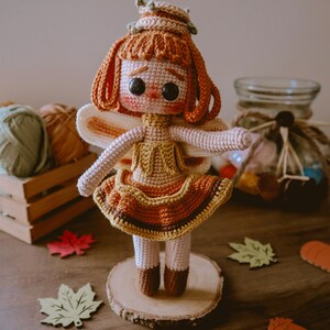 Puede incluir: Una muñeca de hada de crochet con cabello naranja, un vestido marrón y amarillo y alas blancas. La muñeca está de pie sobre una rodaja de madera y rodeada de decoraciones de otoño.