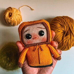 Puede incluir: Un juguete de peluche tejido a crochet con forma de persona que lleva una chaqueta amarilla con capucha. El juguete tiene ojos marrones y una cara rosa. El juguete está siendo sostenido en una mano.
