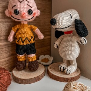 May include: Crocheted amigurumi figures of Charlie Brown and Snoopy. Charlie Brown is wearing a yellow shirt with a black and white zigzag pattern, black trousers, and brown shoes. Snoopy is white with black ears and a red collar. Both figures are standing on wooden rounds.