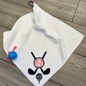 May include: White golf towel with a black and red golf club and ball design. The towel has a waffle texture and a black carabiner clip. A blue golf ball and pink tees are next to the towel.
