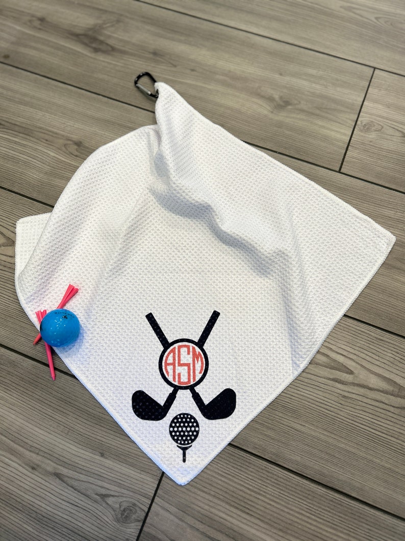 May include: White golf towel with a black and red golf club and ball design. The towel has a waffle texture and a black carabiner clip. A blue golf ball and pink tees are next to the towel.