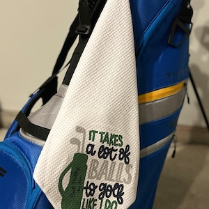 May include: White golf towel with the phrase "IT TAKES a lot of BALLS to golf LIKE I DO" in green, gray, and navy blue. The towel is hanging on a blue golf bag. The towel has a textured surface.