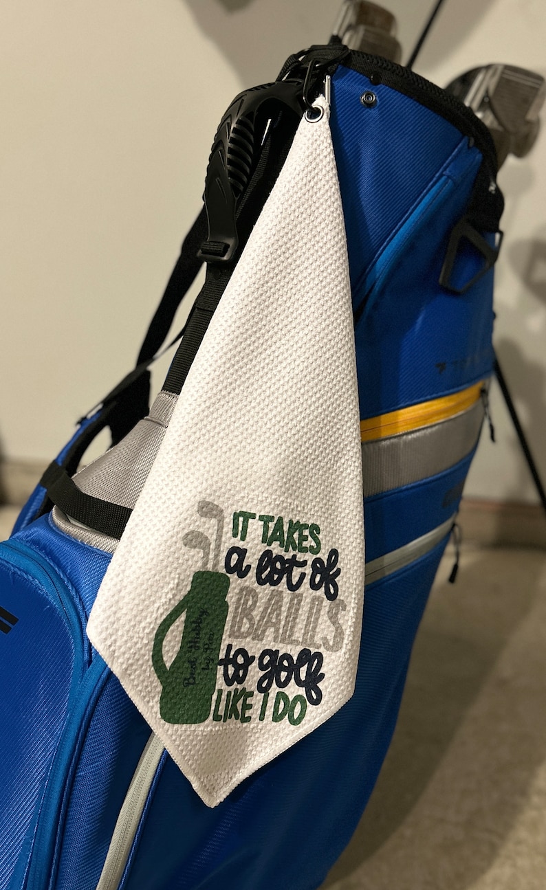May include: White golf towel with the phrase "IT TAKES a lot of BALLS to golf LIKE I DO" in green, gray, and navy blue. The towel is hanging on a blue golf bag. The towel has a textured surface.