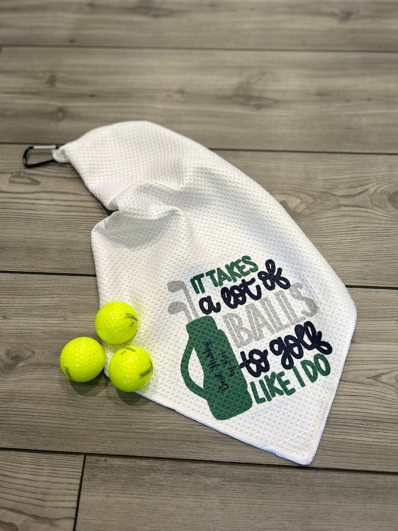 May include: White golf towel with the text "IT TAKES a lot of BALLS to golf LIKE I DO" in gray, green, and navy blue. Three yellow golf balls are next to the towel. The towel has a black carabiner clip.