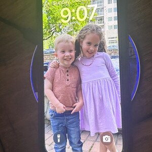 May include: A photo on a phone screen shows two children, a boy and a girl, smiling at the camera. The boy is wearing a pink shirt and blue jeans. The girl is wearing a lavender dress and a pink necklace. The phone screen shows the date "Sunday, December 29" and the time "9:07".