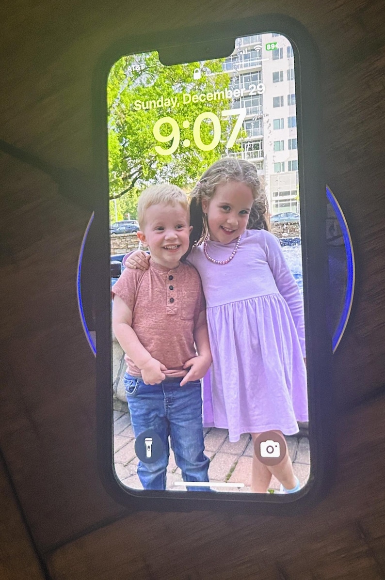 May include: A photo on a phone screen shows two children, a boy and a girl, smiling at the camera. The boy is wearing a pink shirt and blue jeans. The girl is wearing a lavender dress and a pink necklace. The phone screen shows the date "Sunday, December 29" and the time "9:07".