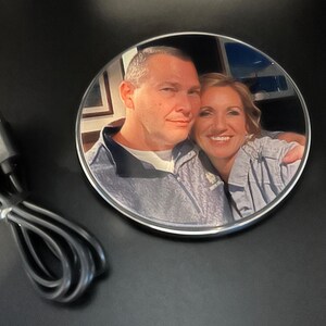 May include: A round, silver wireless charging pad with a photo of a couple printed on the top. The photo shows a man and a woman smiling at the camera. The charging pad is on a black surface.