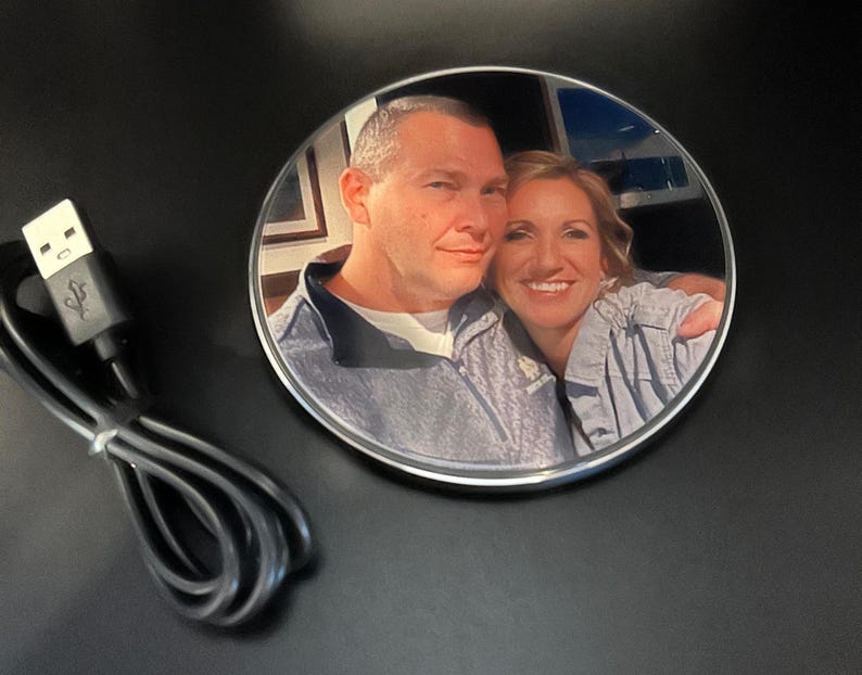 May include: A round, silver wireless charging pad with a photo of a couple printed on the top. The photo shows a man and a woman smiling at the camera. The charging pad is on a black surface.