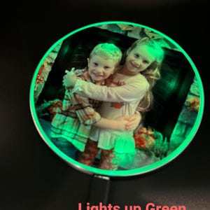 May include: A round, silver-colored photo frame with a green LED light around the edge. The frame contains a photo of two children hugging. The text "Lights up Green when on" is visible below the frame.