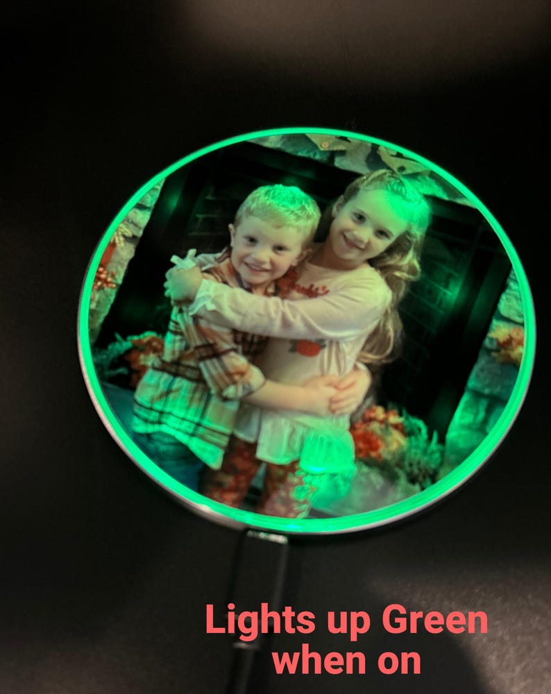 May include: A round, silver-colored photo frame with a green LED light around the edge. The frame contains a photo of two children hugging. The text "Lights up Green when on" is visible below the frame.