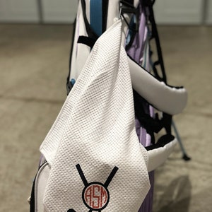 May include: A golf bag with a white towel hanging from it. The towel has a black and red design of crossed golf clubs, a golf ball, and the initials "ASM". The golf bag is white, purple, and blue.