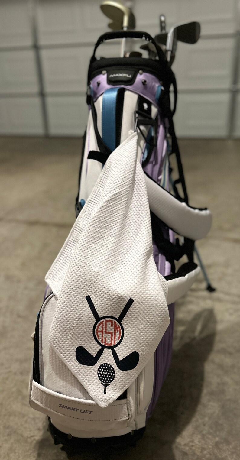 May include: A golf bag with a white towel hanging from it. The towel has a black and red design of crossed golf clubs, a golf ball, and the initials "ASM". The golf bag is white, purple, and blue.